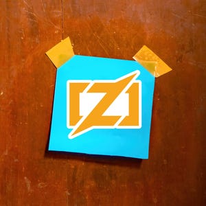 Puede incluir: Un cuadrado azul con un logotipo naranja y blanco, pegado a una superficie de madera con cinta adhesiva. El logotipo presenta una letra "Z" naranja estilizada dentro de un rect&aacute;ngulo blanco, con un rayo naranja.