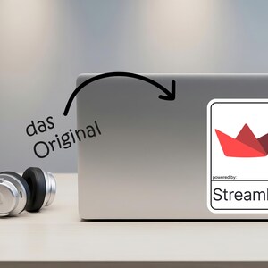 K&ouml;nnte beinhalten: Ein Laptop mit einem Aufkleber mit einem roten Papierboot-Design und dem Text "powered by Streamlit". Die Worte "das Original" sind auf dem Laptop geschrieben. Silberne Kopfh&ouml;rer liegen auf dem Tisch.