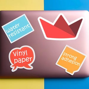 Op de afbeelding: Laptop bedekt met stickers. Stickers zijn onder meer een rode origami-kroon, een blauwe rechthoek met "water resistant", een rode tekstballon met "vinyl paper" en een oranje vierkant met "strong adhesion".