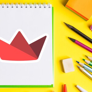 Op de afbeelding: Een witte notitieblok met een rode papieren kroonsticker. De afbeelding bevat ook potloden, een gum en een puntenslijper op een gele achtergrond. Kantoorartikelen voor school of kantoor.