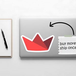 Op de afbeelding: Een rode papieren bootsticker op een laptop, met een briefje met de tekst "buy more, ship once". Een zwarte pen en een wit notitieblok bevinden zich aan de linkerkant van de afbeelding.