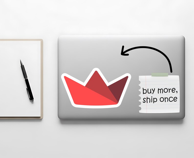 Op de afbeelding: Een rode papieren bootsticker op een laptop, met een briefje met de tekst "buy more, ship once". Een zwarte pen en een wit notitieblok bevinden zich aan de linkerkant van de afbeelding.