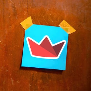 Op de afbeelding: Een vierkante, lichtblauwe post-it met een rood-wit kroonontwerp, bevestigd op een houten oppervlak met twee stukjes gele tape. De kroon heeft een geometrische vorm.