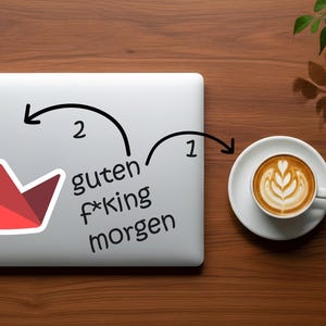 Op de afbeelding: Een laptop met een rood-witte kroonsticker en de Duitse zin "guten f*king morgen" staat naast een kop koffie met latte art op een houten oppervlak. Een takje bladeren bevindt zich in de rechterbovenhoek.