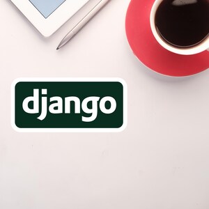 Może przedstawiać: Biała powierzchnia z zielono-białą naklejką "django", srebrnym rysikiem, białym tabletem i czerwoną filiżanką kawy z czarną kawą.