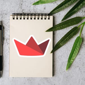 Op de afbeelding: Een rode en kastanjebruine kroonsticker met een witte rand staat op een cr&egrave;mekleurig notitieblok. Een zilveren pen met een zwarte grip staat links van het notitieblok. Groene bladeren bevinden zich in de rechterbovenhoek.