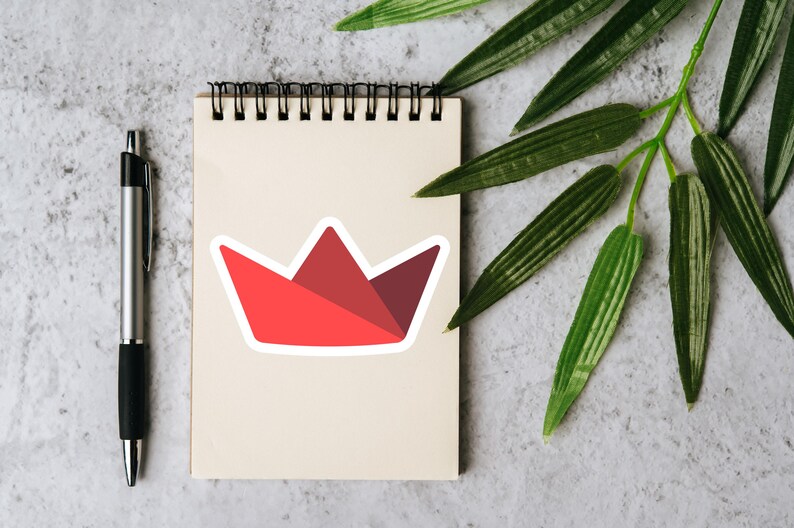 Op de afbeelding: Een rode en kastanjebruine kroonsticker met een witte rand staat op een cr&egrave;mekleurig notitieblok. Een zilveren pen met een zwarte grip staat links van het notitieblok. Groene bladeren bevinden zich in de rechterbovenhoek.