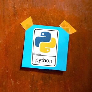 May include: A blue square with the Python logo, featuring a blue and yellow snake-like design, is taped to a wooden surface. The text "powered by: python" is below the logo. Two pieces of yellow tape secure the square.