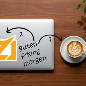 Puede incluir: Un port&aacute;til con una pegatina con un dise&ntilde;o estilizado naranja y blanco y el texto "guten f*king morgen". Una taza de caf&eacute; con arte latte est&aacute; sobre un platillo a la derecha. La escena est&aacute; sobre una superficie de madera.