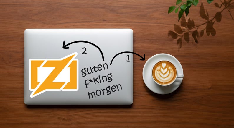 Puede incluir: Un port&aacute;til con una pegatina con un dise&ntilde;o estilizado naranja y blanco y el texto "guten f*king morgen". Una taza de caf&eacute; con arte latte est&aacute; sobre un platillo a la derecha. La escena est&aacute; sobre una superficie de madera.