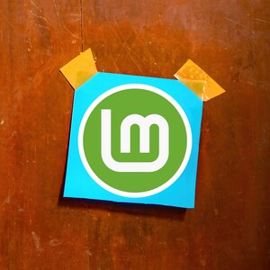 May include: A blue square of paper with the Linux Mint logo, a white stylized "LM" inside a green circle, is taped to a wooden surface with two pieces of yellow tape. The wood has a brown color.