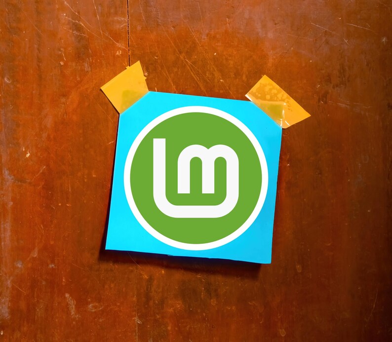 May include: A blue square of paper with the Linux Mint logo, a white stylized "LM" inside a green circle, is taped to a wooden surface with two pieces of yellow tape. The wood has a brown color.