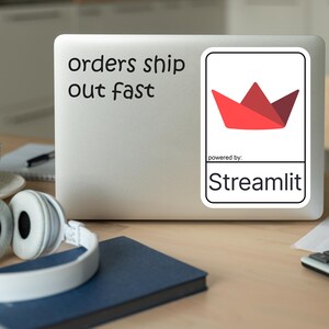 K&ouml;nnte beinhalten: Ein Laptop mit dem Text "orders ship out fast" und einem Aufkleber mit einem roten Papierkronen-Design. Der Aufkleber ist mit "powered by: Streamlit" beschriftet. Wei&szlig;e Kopfh&ouml;rer, eine Tasse und ein Notizbuch liegen auf dem Holztisch.