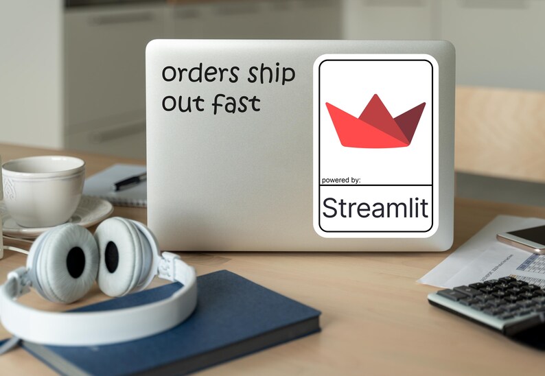 K&ouml;nnte beinhalten: Ein Laptop mit dem Text "orders ship out fast" und einem Aufkleber mit einem roten Papierkronen-Design. Der Aufkleber ist mit "powered by: Streamlit" beschriftet. Wei&szlig;e Kopfh&ouml;rer, eine Tasse und ein Notizbuch liegen auf dem Holztisch.