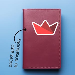 Op de afbeelding: Een bordeauxrood notitieboek met een rood-witte kroonsticker. De sticker heeft een geometrisch ontwerp met een witte omtrek. De tekst "sticks also to notebooks" staat naast het notitieboek.