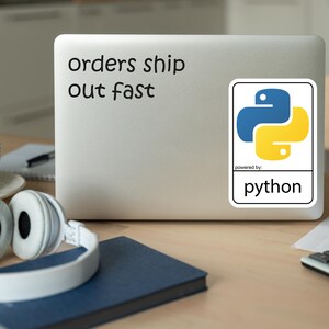 May include: A laptop with a sticker featuring the Python logo in blue and yellow. The text "orders ship out fast" is above the sticker. Headphones, a cup, and a notebook are on the desk.