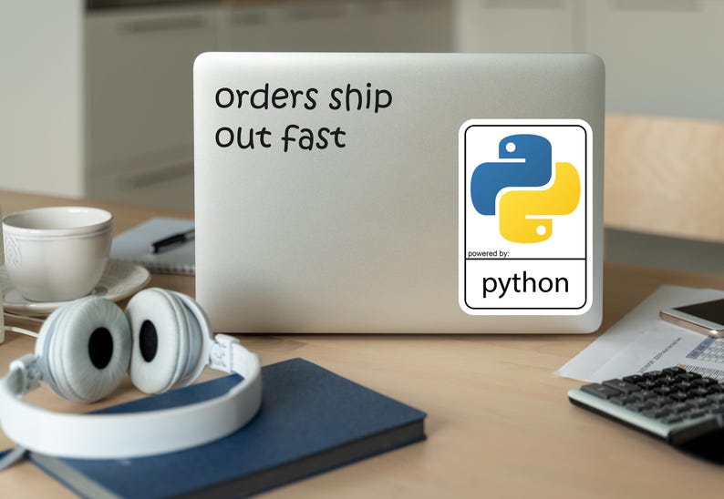 May include: A laptop with a sticker featuring the Python logo in blue and yellow. The text "orders ship out fast" is above the sticker. Headphones, a cup, and a notebook are on the desk.