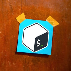 May include: A blue sticker with a white and black cube logo, featuring a dollar sign and a green underscore. The sticker is affixed to a wooden surface with two pieces of yellow tape.