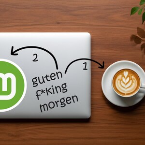 May include: A laptop with a green and white logo and the words "guten f*king morgen" is next to a cup of coffee with latte art on a wooden surface. A sprig of leaves is in the upper right corner.