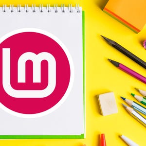May include: A spiral-bound notepad with a white page featuring a magenta circle with a white "lm" logo. Pencils, an eraser, and a pencil sharpener are scattered on a bright yellow surface.