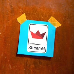 K&ouml;nnte beinhalten: Ein hellblaues Quadrat mit einem wei&szlig;en Rechteck, das ein rotes und kastanienbraunes Papierboot-Logo und den Text "powered by: Streamlit" enth&auml;lt. Das Quadrat ist mit gelbem Klebeband auf einer Holzoberfl&auml;che befestigt.