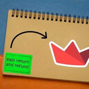 Op de afbeelding: Een spiraalgebonden notitieboek met een rode papieren kroonsticker en een groene vierkante sticker met de tekst "easy return and refund". Een houten pen met gouden accenten staat links.