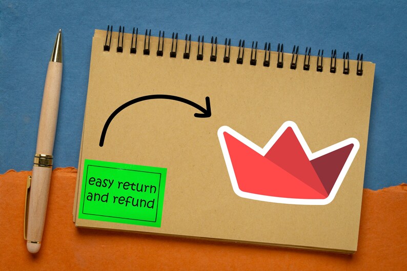 Op de afbeelding: Een spiraalgebonden notitieboek met een rode papieren kroonsticker en een groene vierkante sticker met de tekst "easy return and refund". Een houten pen met gouden accenten staat links.