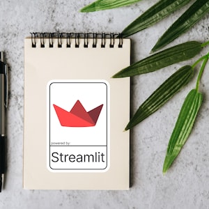 K&ouml;nnte beinhalten: Ein Notizblock mit schwarzer Spiralbindung, ein silberner Stift und ein Aufkleber mit einem roten Papierboot-Design. Der Aufkleber tr&auml;gt den Text "powered by: Streamlit" in schwarzer Schrift. Gr&uuml;ne Bl&auml;tter sind im Hintergrund.