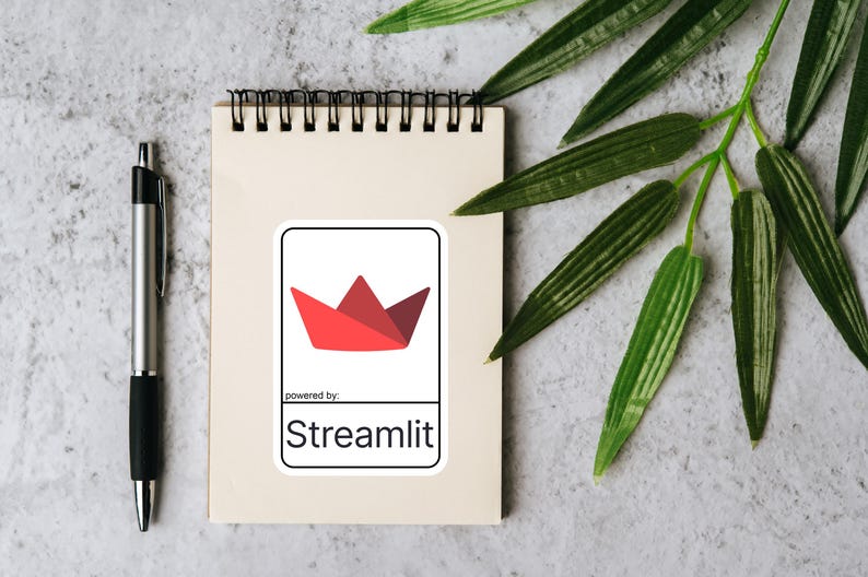 K&ouml;nnte beinhalten: Ein Notizblock mit schwarzer Spiralbindung, ein silberner Stift und ein Aufkleber mit einem roten Papierboot-Design. Der Aufkleber tr&auml;gt den Text "powered by: Streamlit" in schwarzer Schrift. Gr&uuml;ne Bl&auml;tter sind im Hintergrund.