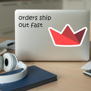 Op de afbeelding: Een laptop met een sticker met de tekst "orders ship out fast" en een rode kroongrafiek. De laptop staat op een houten bureau met een koptelefoon, een koffiekopje, een notitieboek en een rekenmachine.