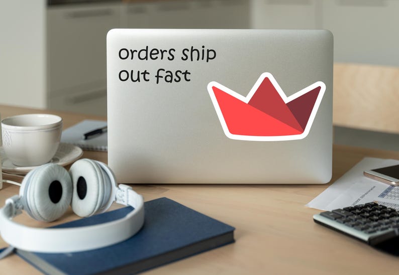 Op de afbeelding: Een laptop met een sticker met de tekst "orders ship out fast" en een rode kroongrafiek. De laptop staat op een houten bureau met een koptelefoon, een koffiekopje, een notitieboek en een rekenmachine.