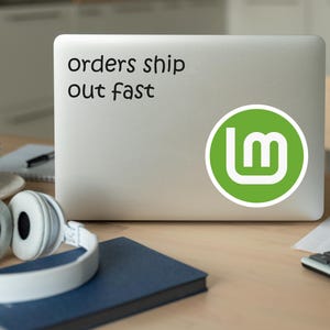 May include: A laptop with a sticker that says "orders ship out fast" and a green logo. White headphones, a cup, a notebook, and a calculator are on the wooden desk.