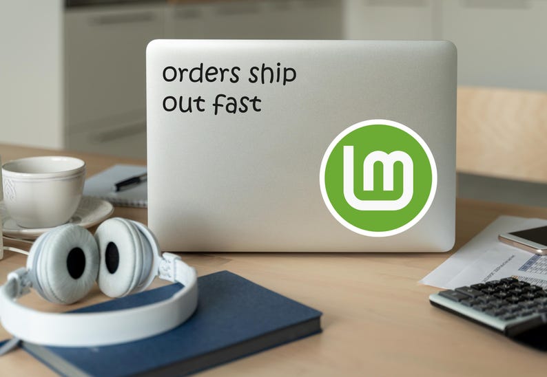 May include: A laptop with a sticker that says "orders ship out fast" and a green logo. White headphones, a cup, a notebook, and a calculator are on the wooden desk.