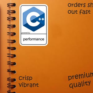 May include: An orange spiral-bound notebook with a black pen and a sticker featuring a blue and white C++ logo. The sticker reads "powered by: performance." Additional text includes "Crisp Vibrant" and "premium quality."
