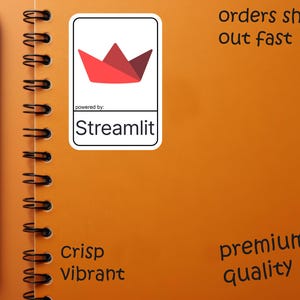 K&ouml;nnte beinhalten: Ein orangefarbenes Spiralnotizbuch mit einem schwarzen Stift und einem wei&szlig;en Aufkleber mit einem roten Papierboot-Design und dem Text "Streamlit". Das Notizbuch hat auch die W&ouml;rter "Crisp Vibrant" und "premium quality".