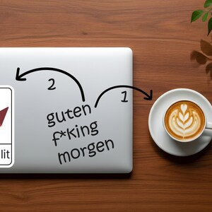K&ouml;nnte beinhalten: Ein Laptop mit einem Streamlit-Aufkleber und den Worten "guten f*king morgen" darauf geschrieben. Rechts steht eine Tasse Kaffee mit Latte Art. Das Bild befindet sich auf einer Holzoberfl&auml;che.