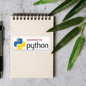 May include: A notepad with a spiral binding, a silver pen, and a sticker that says "powered by python" with a blue and yellow logo. Green leaves are in the background. The notepad is blank.