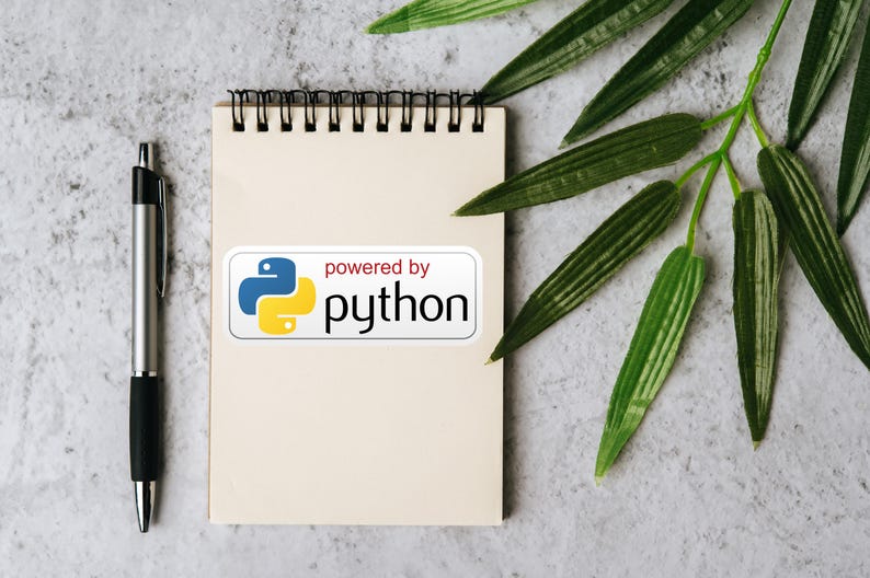 May include: A notepad with a spiral binding, a silver pen, and a sticker that says "powered by python" with a blue and yellow logo. Green leaves are in the background. The notepad is blank.