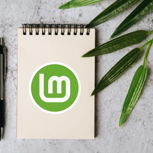May include: A spiral-bound notepad with a green and white logo, a silver pen, and green leaves on a textured gray surface. The logo is a green circle with a white "lm" design. The pen is silver with a black grip.