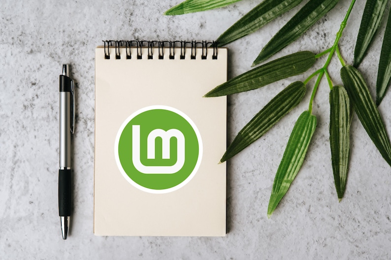 May include: A spiral-bound notepad with a green and white logo, a silver pen, and green leaves on a textured gray surface. The logo is a green circle with a white "lm" design. The pen is silver with a black grip.
