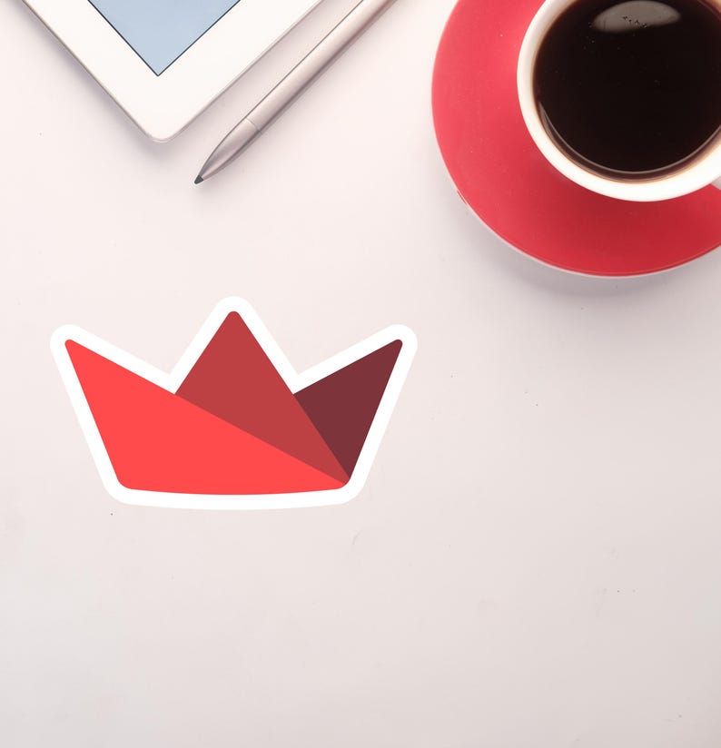 Op de afbeelding: Een rode papieren kroonsticker met een witte rand. De sticker bevindt zich op een wit oppervlak, met een tablet, stylus en een kopje koffie op een rode schotel op de achtergrond.