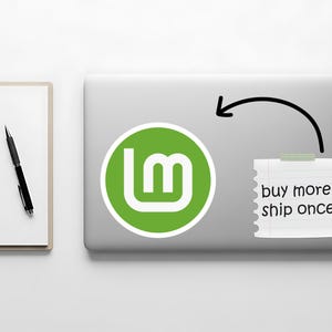 May include: A laptop with a green and white logo sticker and a sticky note that says "buy more, ship once". A black pen and a white notepad are on the left.