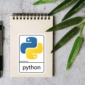 May include: A notepad with a Python sticker, a silver pen, and green leaves on a textured gray surface. The sticker features the Python logo in blue and yellow, with the text "powered by: python".