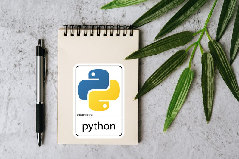 May include: A notepad with a Python sticker, a silver pen, and green leaves on a textured gray surface. The sticker features the Python logo in blue and yellow, with the text "powered by: python".