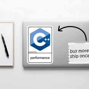 May include: A laptop with a sticker featuring the C++ logo, a blue and white hexagon design. A notepad with the text "buy more, ship once" is taped to the laptop. A pen and notepad are on the left.