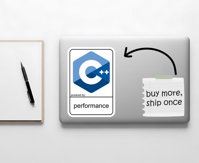 May include: A laptop with a sticker featuring the C++ logo, a blue and white hexagon design. A notepad with the text "buy more, ship once" is taped to the laptop. A pen and notepad are on the left.