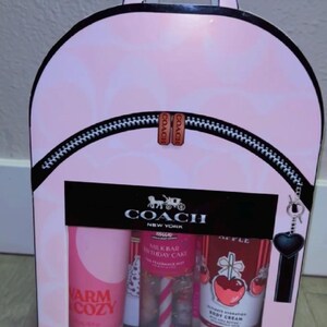 May include: A Coach gift set packaged in a light pink backpack-shaped box with a repeating logo pattern. The box features a black zipper detail, a Coach logo, and a clear window displaying three body care products: Warm Cozy, Milk Bar Birthday Cake fragrance mist, and an apple-themed body cream.