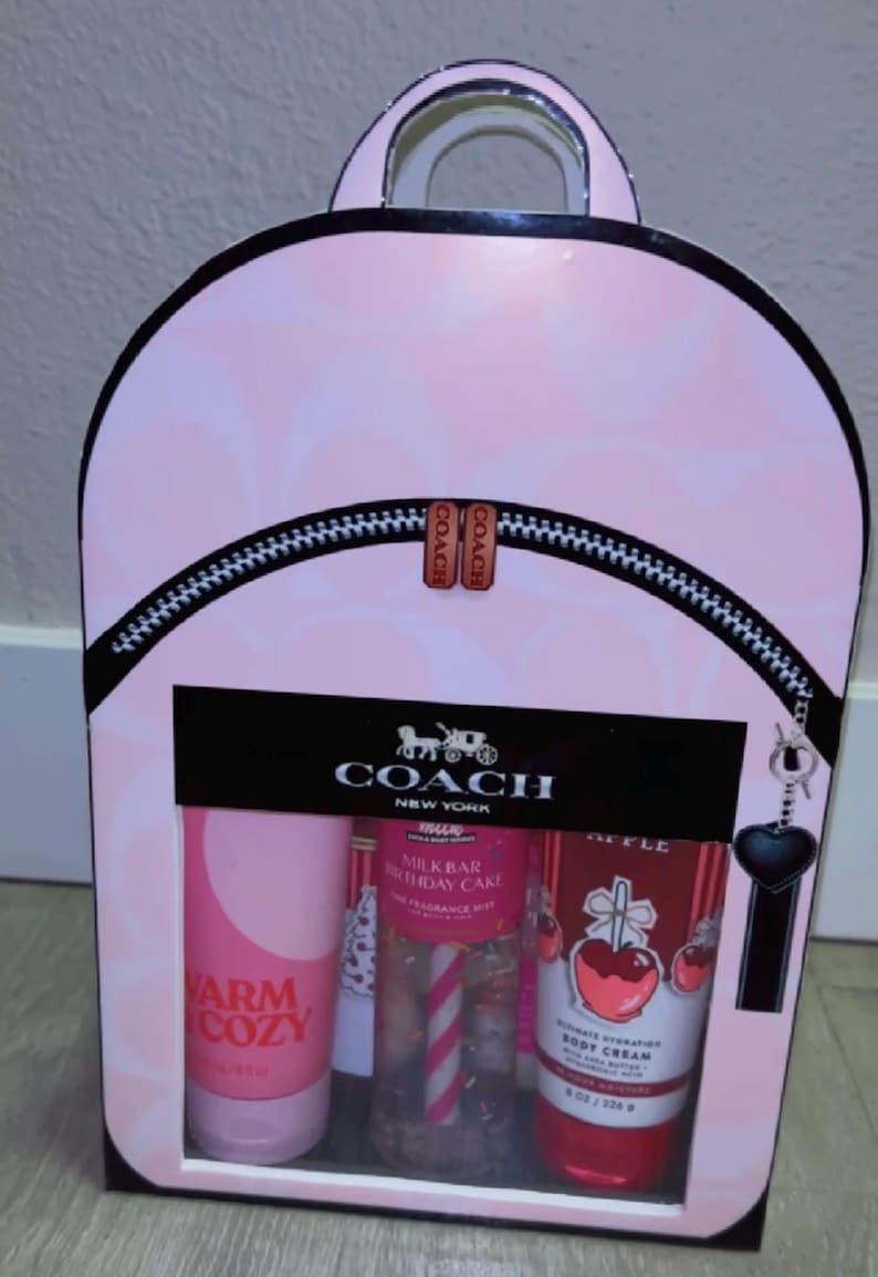 May include: A Coach gift set packaged in a light pink backpack-shaped box with a repeating logo pattern. The box features a black zipper detail, a Coach logo, and a clear window displaying three body care products: Warm Cozy, Milk Bar Birthday Cake fragrance mist, and an apple-themed body cream.