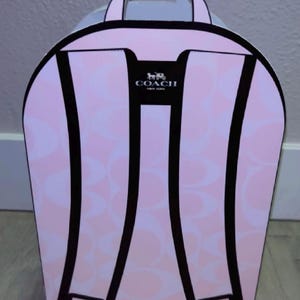 May include: A light pink cardboard box shaped like a backpack with a repeating Coach logo pattern. The front features a graphic illustration of black backpack straps and a top handle. The Coach New York logo is printed in black at the top center.