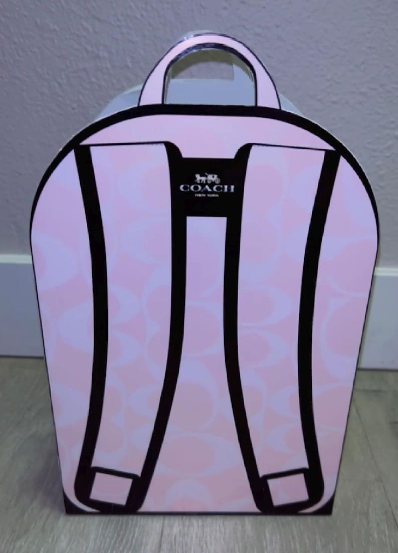May include: A light pink cardboard box shaped like a backpack with a repeating Coach logo pattern. The front features a graphic illustration of black backpack straps and a top handle. The Coach New York logo is printed in black at the top center.